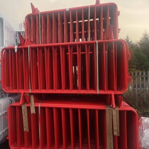 Stack of vibrant red fixed-leg crowd control barriers, 2.3m x 1.1m, neatly bundled and stored outdoors on a construction yard with green straps and nearby white barriers in the background.