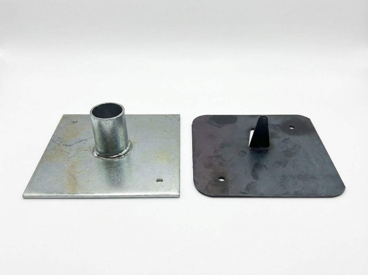 Side-by-side comparison of two scaffold base plates: galvanised steel with central tubular spigot socket on the left, and heavy-duty black painted plate with pointed locator pin on the right, both square with fixing holes.