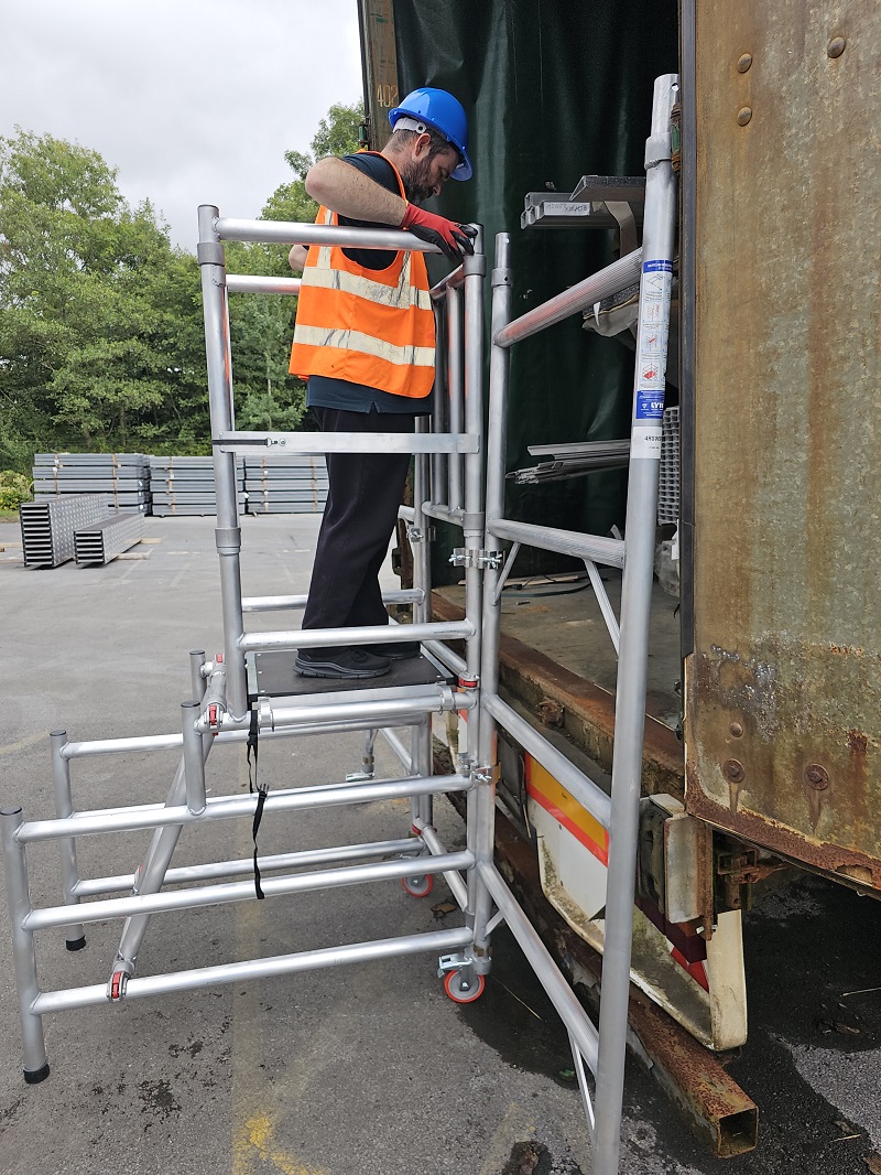 Side view of worker in high-vis and hard hat on aluminium mobile podium platform adjusting setup at open truck rear doors.
