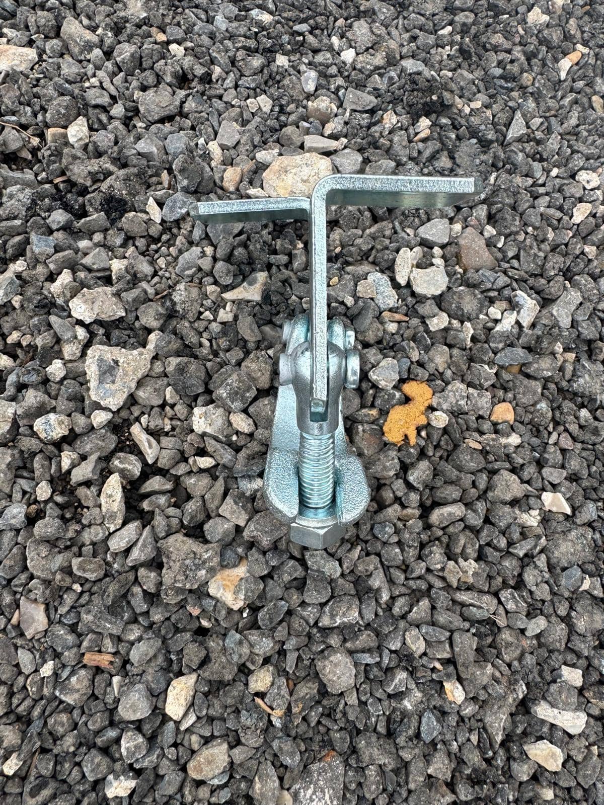Board retaining clamp positioned on gravel surface, highlighting the L-shaped flange and bolt mechanism for secure scaffold board retention.