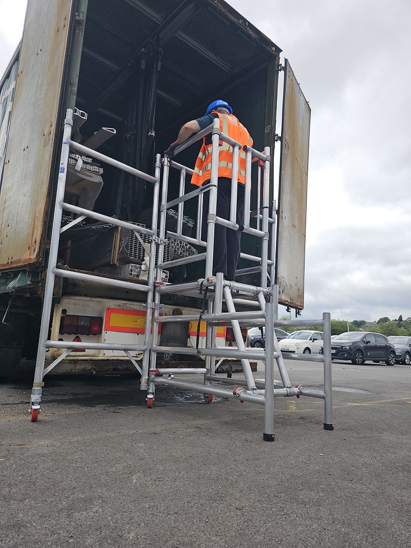 Worker in safety gear standing on platform of mobile aluminium podiumaccessing the interior of a lorry trailer bed.